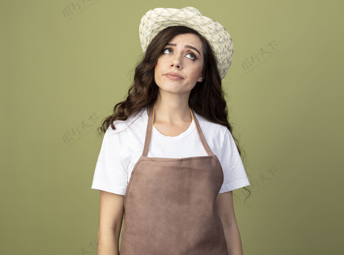 帽子穿着制服 戴着园艺帽 不自信的年轻女园丁看着橄榄绿墙上孤立的一面表情脸橄榄