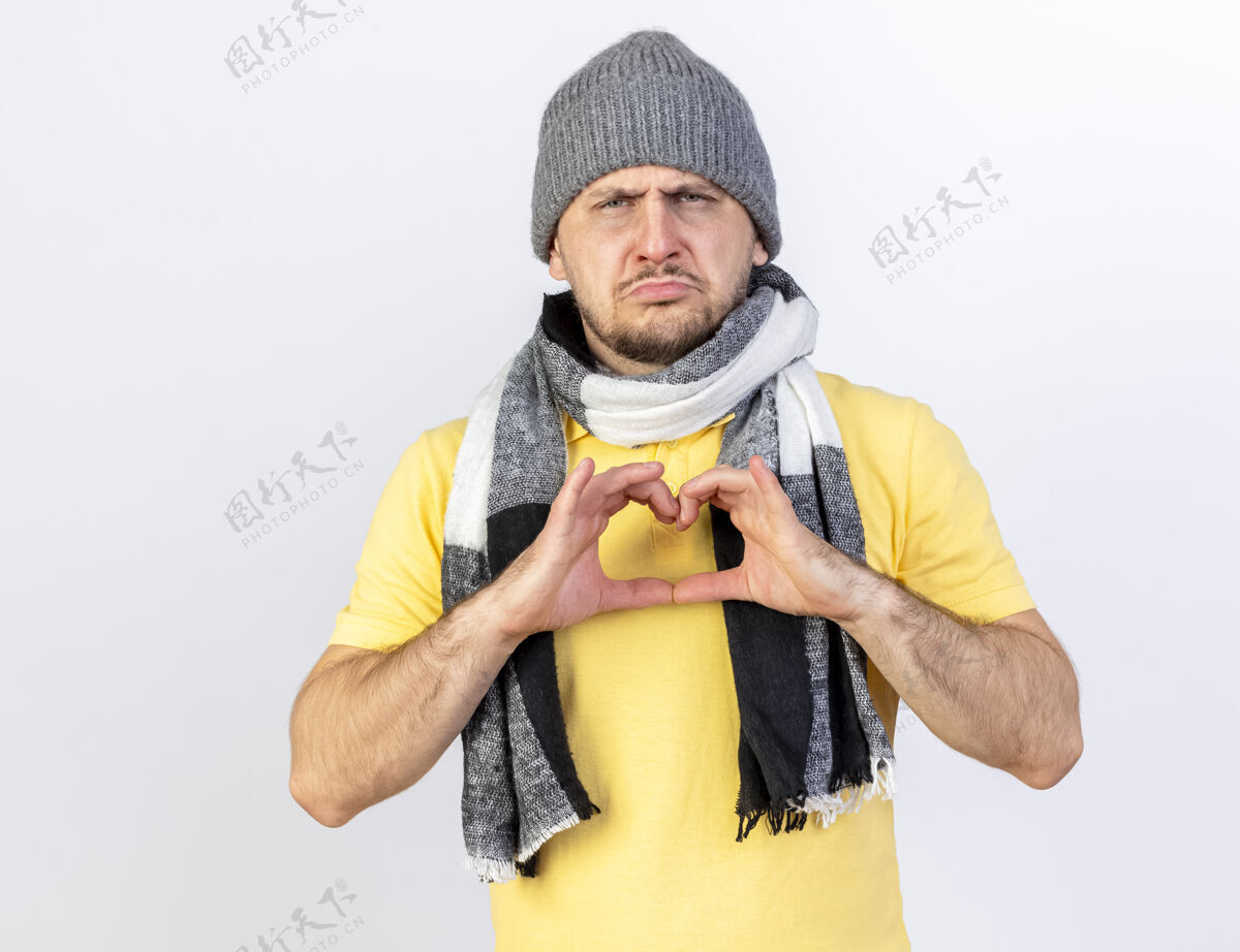 冬天悲伤的年轻金发病男子戴着冬天的帽子和围巾手势心脏标志隔离在白墙上表情姿势帽子