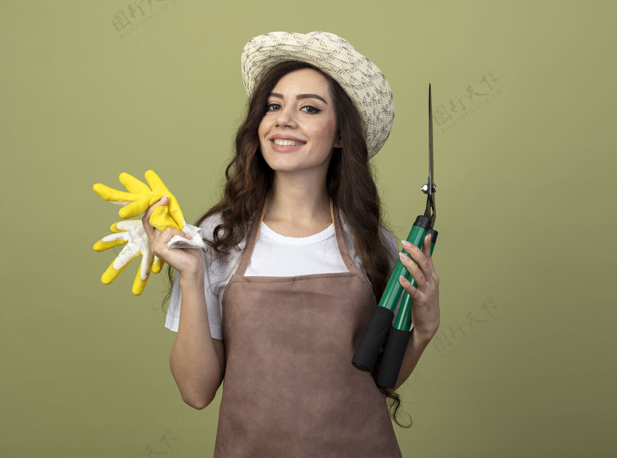 手套微笑的年轻女园丁穿着制服 戴着园艺帽 手持修剪器和手套 隔离在橄榄绿的墙上姿势人微笑
