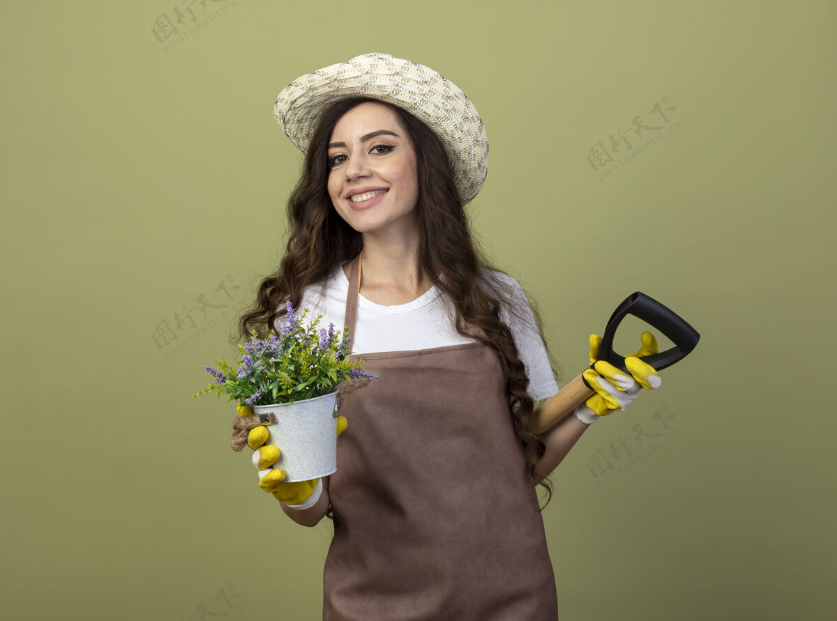 表情身着制服 戴着园艺帽和手套 面带微笑的年轻女园丁背着花盆和铁锹 孤零零地站在橄榄绿的墙上帽子人黑桃