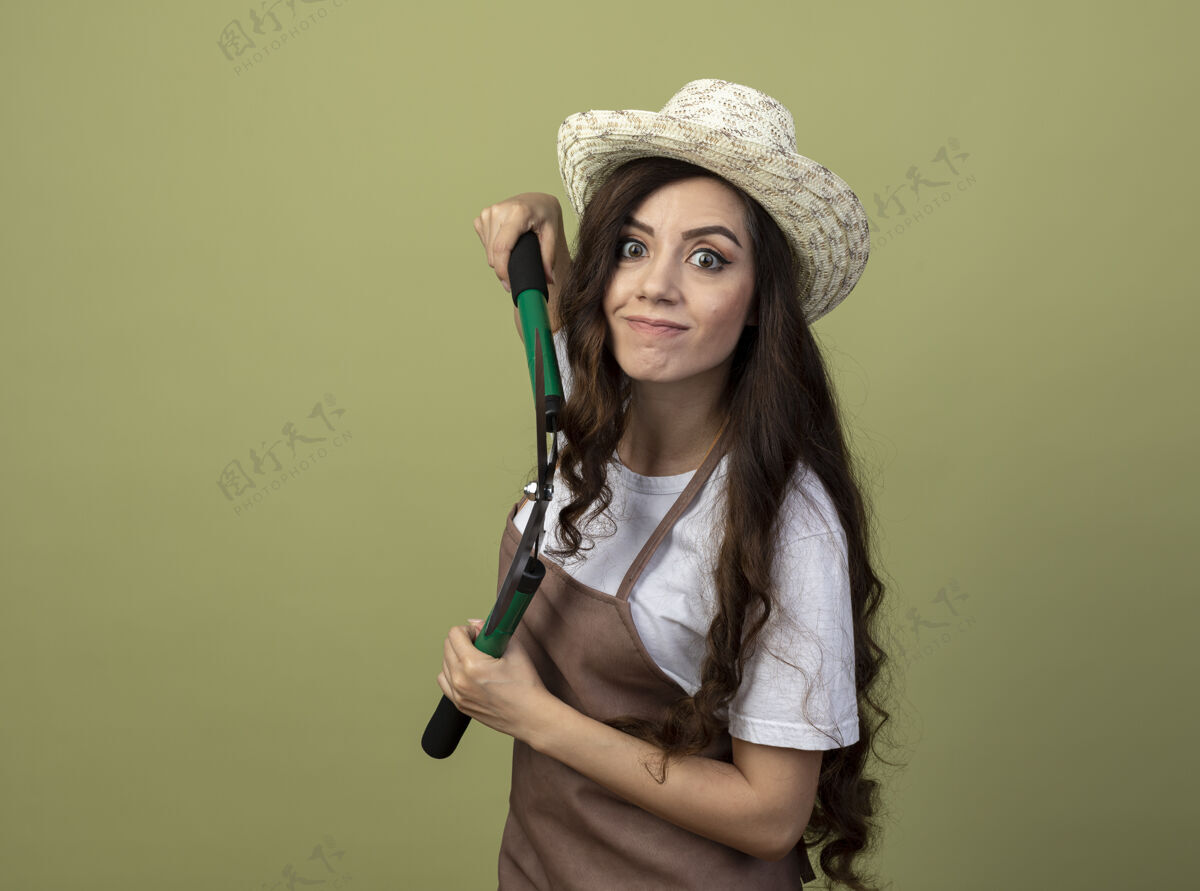 园艺年轻的女园丁穿着制服 戴着园艺帽 站在一旁 拿着园艺剪 隔离在橄榄绿的墙上花园人立场