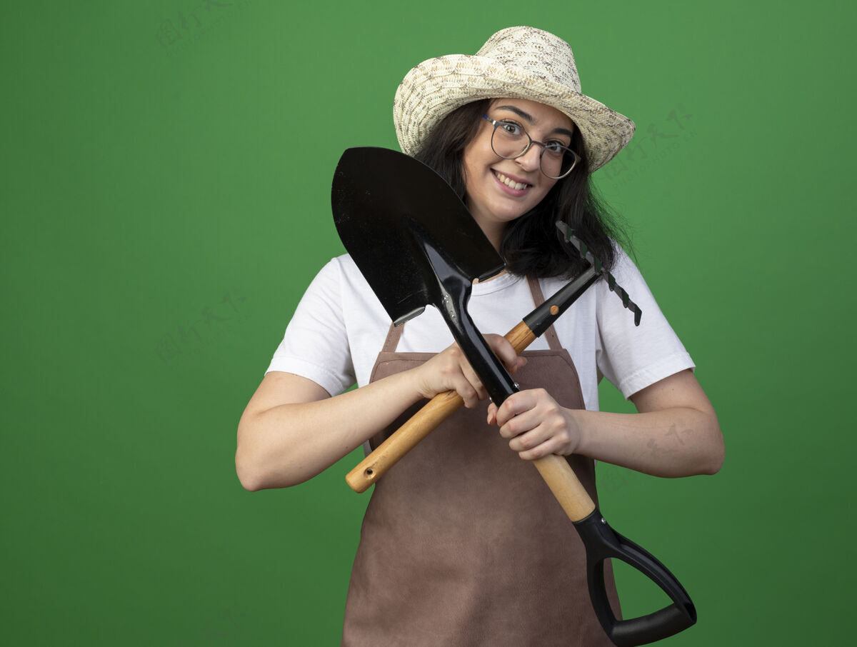 耙子兴奋的年轻黑发女园丁戴着眼镜和制服 戴着园艺帽 把铲子和耙子孤立在绿色的墙上人姿势持有