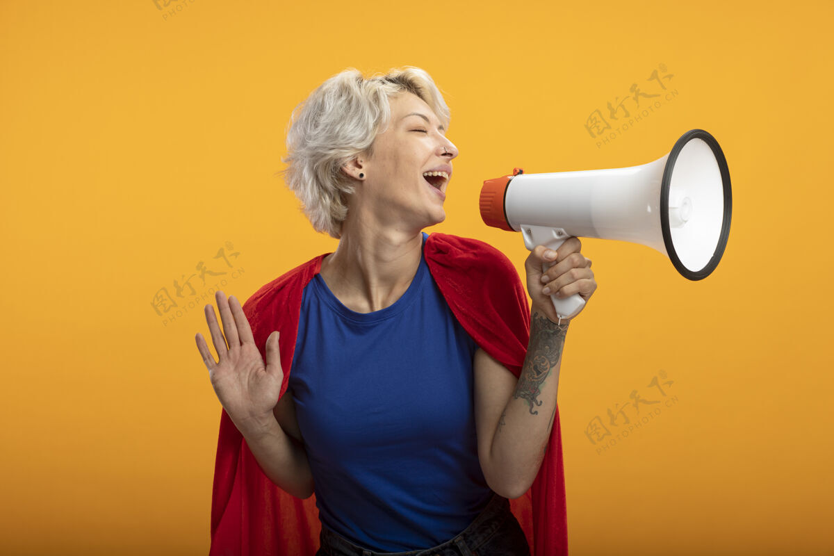 演说家身着红色斗篷的快乐女超人在橙色的墙上举着一个孤立的扩音器超级英雄人女人