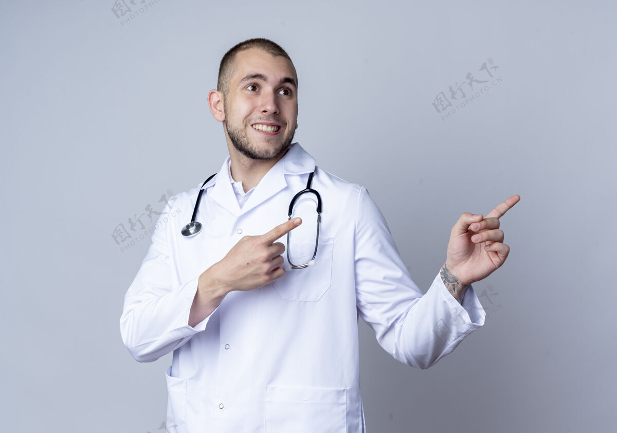年轻人面带微笑的年轻男医生 穿着医用长袍 脖子上戴着听诊器 指着隔离在白墙上的一边看穿着医学脖子
