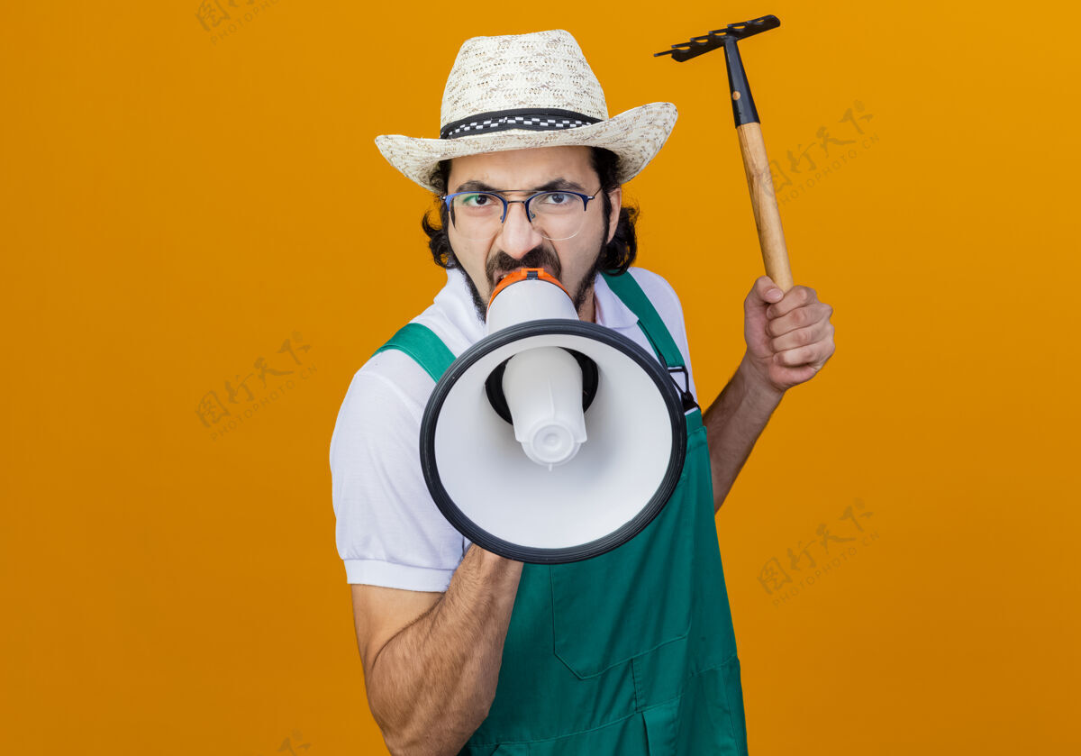 抱着年轻的留着胡须的园丁 穿着连体衣 戴着帽子 手里拿着小耙子 站在橘红色的墙上对着扩音器大喊大叫 表情咄咄逼人人扩音器姿势