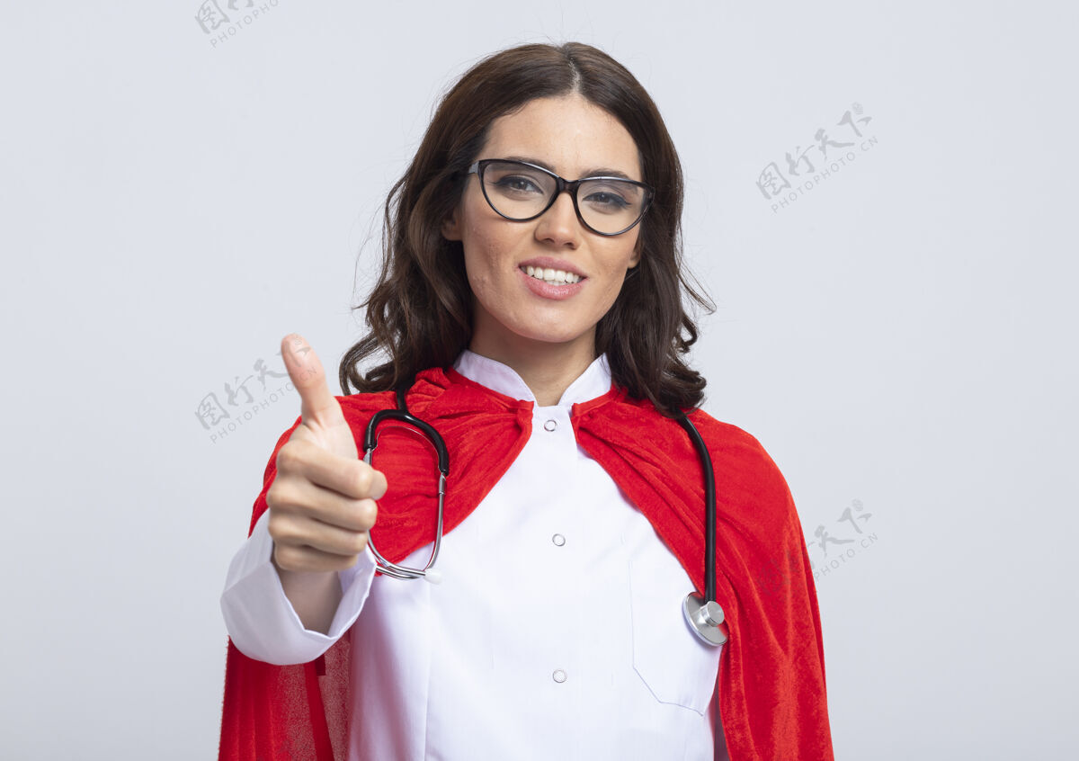 姿势微笑的女超人穿着医生制服 戴着红色斗篷和听诊器 戴着眼镜 竖起大拇指孤立地站在白色的墙上光学拇指听诊器