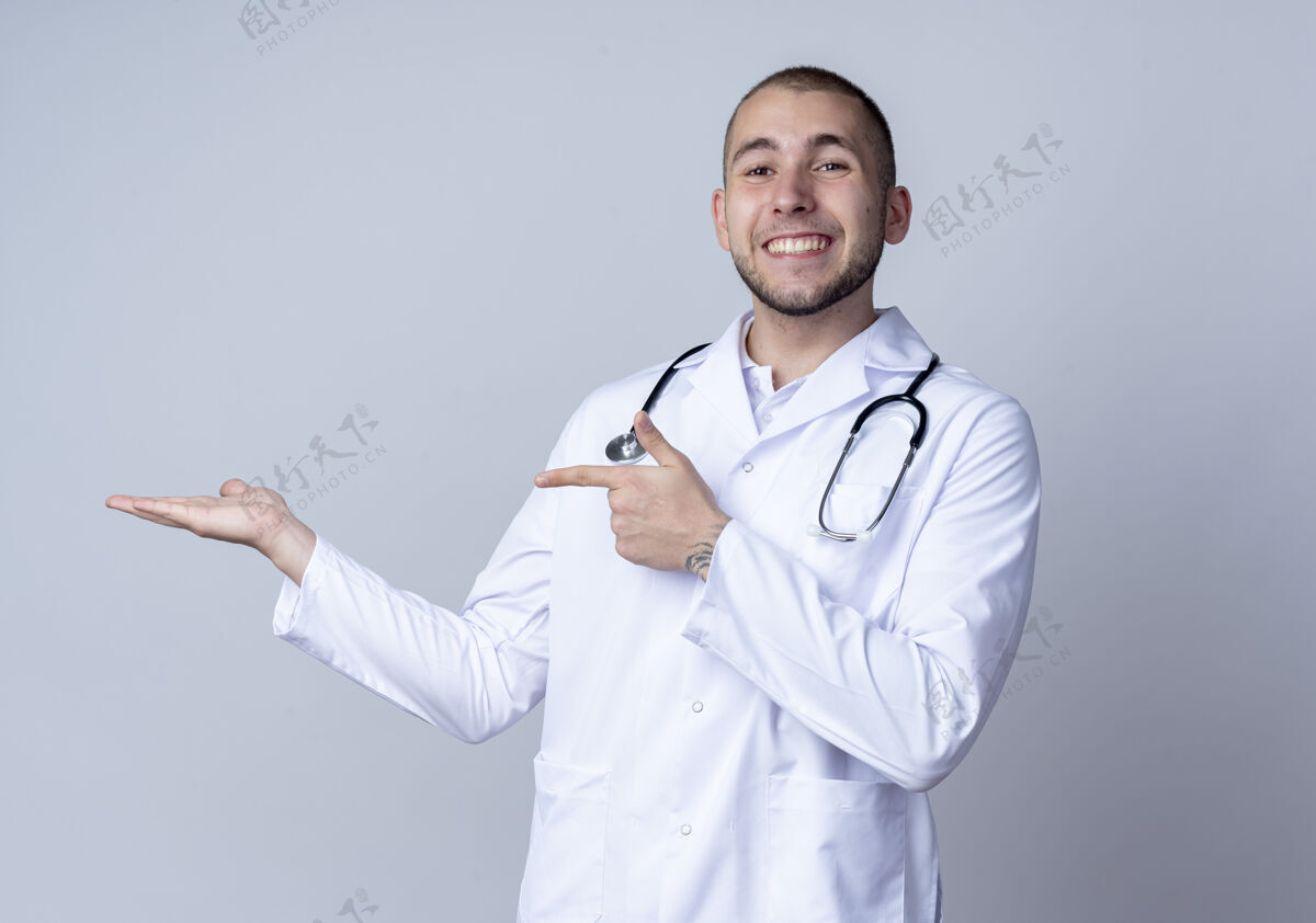 手面带微笑的年轻男医生 身穿医用长袍 脖子上戴着听诊器 伸出空手 指着隔离在白墙上的那只手男性公民听诊器