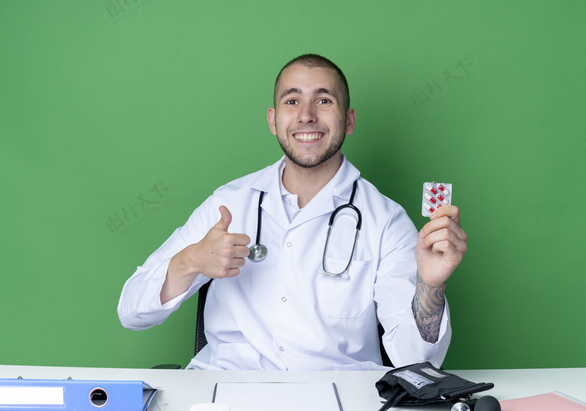 医生面带微笑的年轻男医生穿着医用长袍 听诊器坐在办公桌旁 手里拿着一包胶囊 在绿色的墙上孤立地竖起大拇指工具胶囊表情