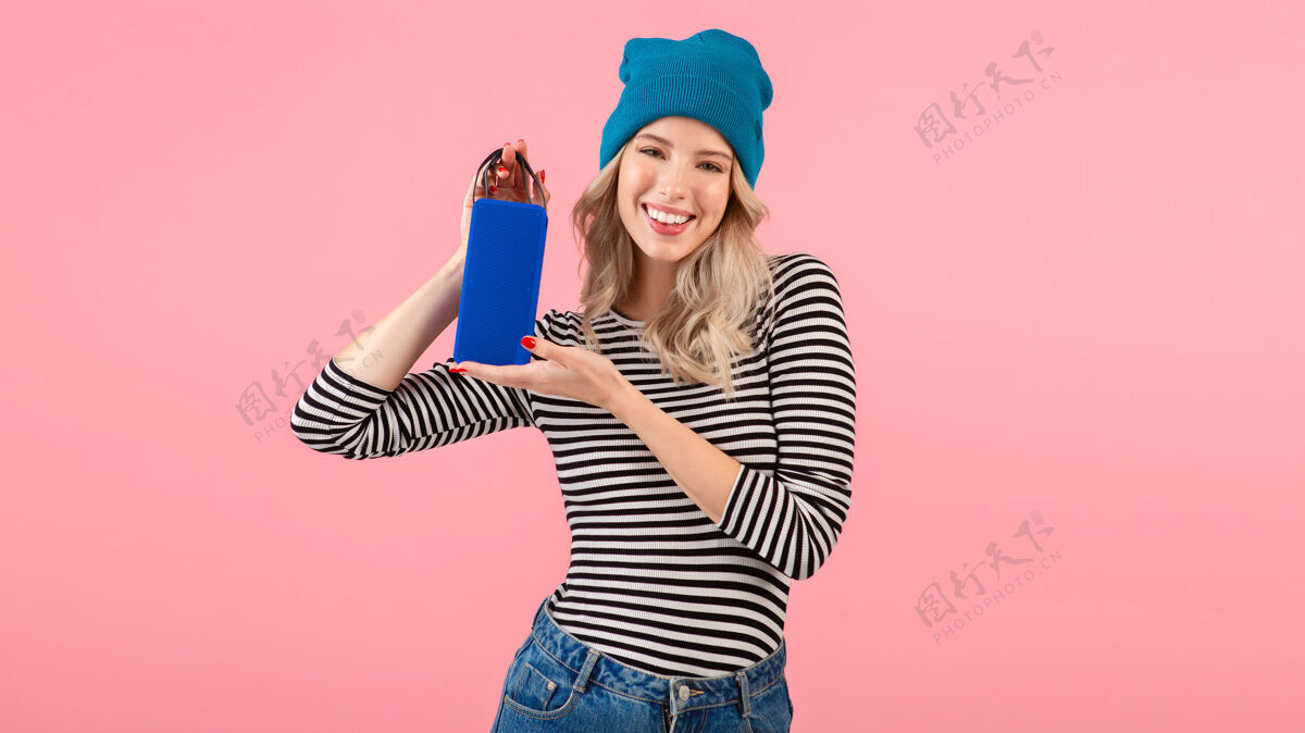 女性年轻女子手持无线扬声器听音乐穿着条纹衬衫和蓝色帽子微笑着摆出粉红色的姿势积极快乐乐趣
