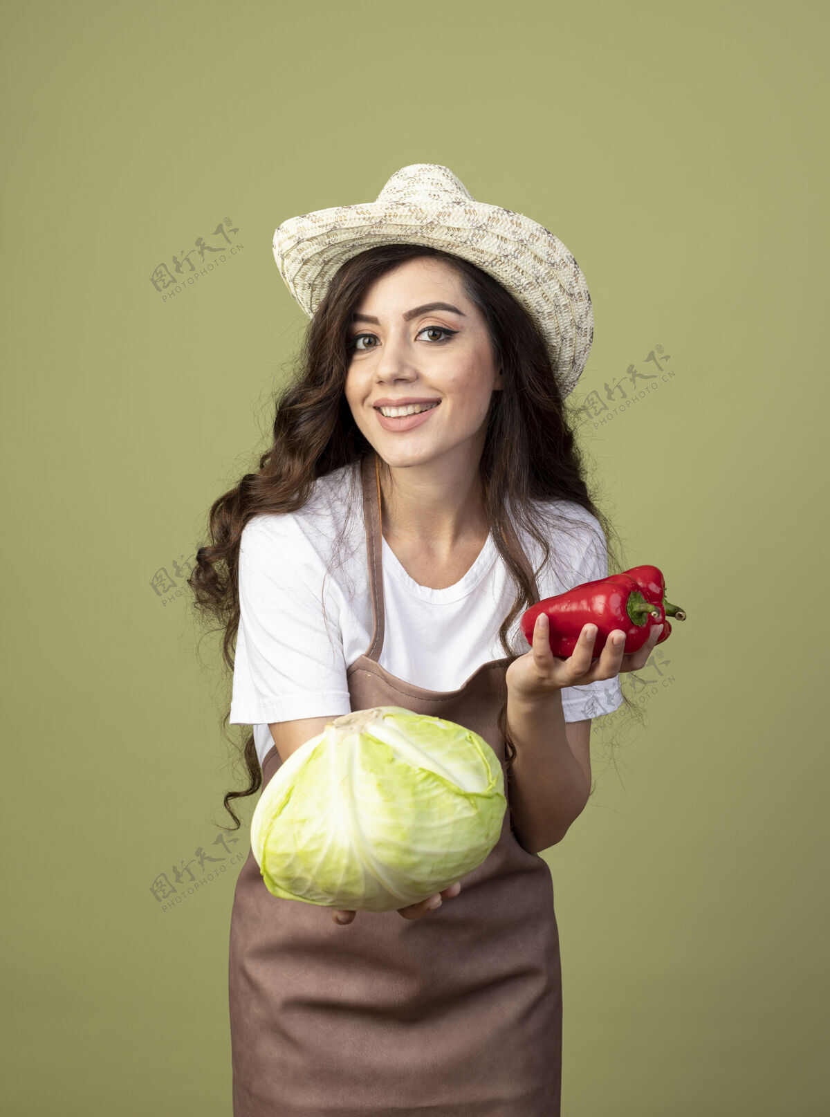 帽子身着制服 头戴园艺帽 面带微笑的年轻女园丁把卷心菜和红辣椒隔离在橄榄绿的墙上卷心菜姿势市民