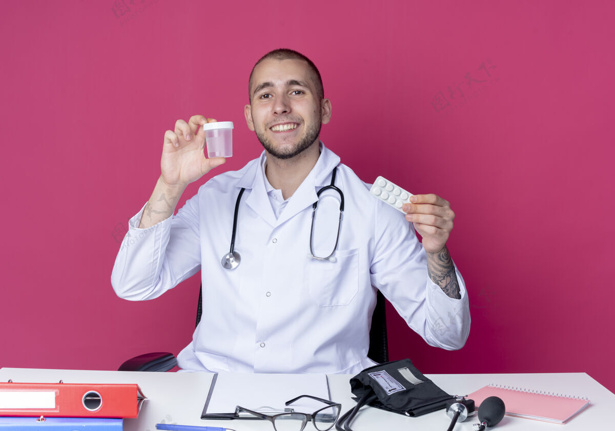 拿着面带微笑的年轻男医生穿着医用长袍 听诊器坐在办公桌旁 手里拿着工作工具 手里拿着医用烧杯和隔离在粉红色墙上的一包药片表情药片工作