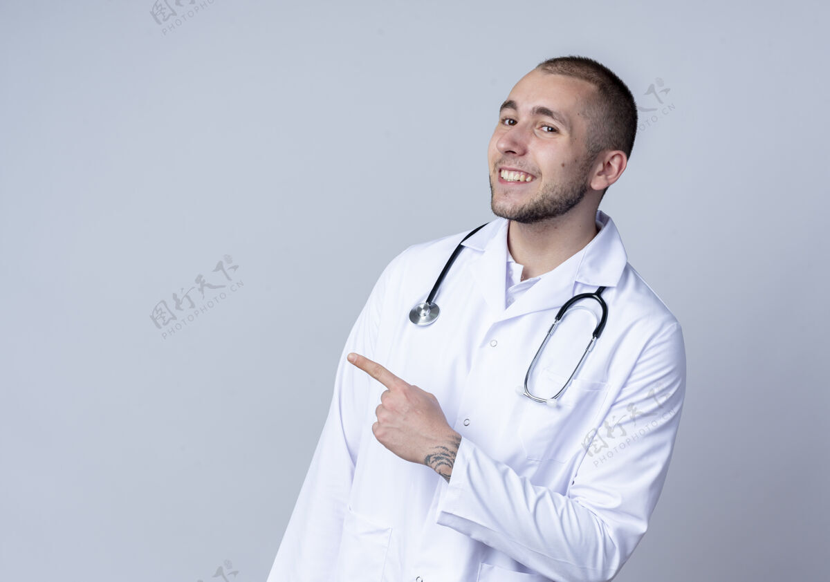 人面带微笑的年轻男医生 身穿医用长袍 脖子上戴着听诊器 指着隔离在白墙上的一侧男性脸人