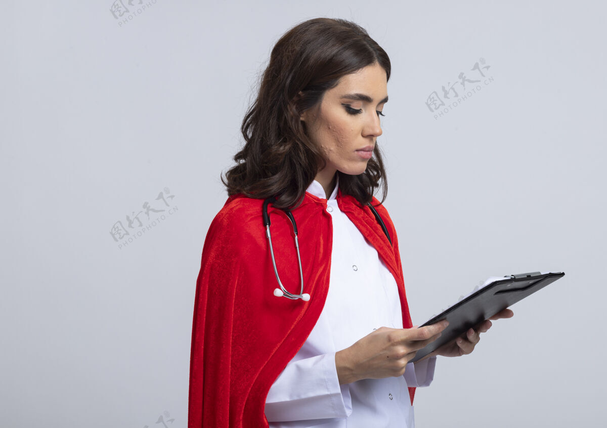 举行自信的女超人穿着医生制服 披着红色斗篷 手持听诊器 看着隔离在白墙上的剪贴板表情剪贴板制服
