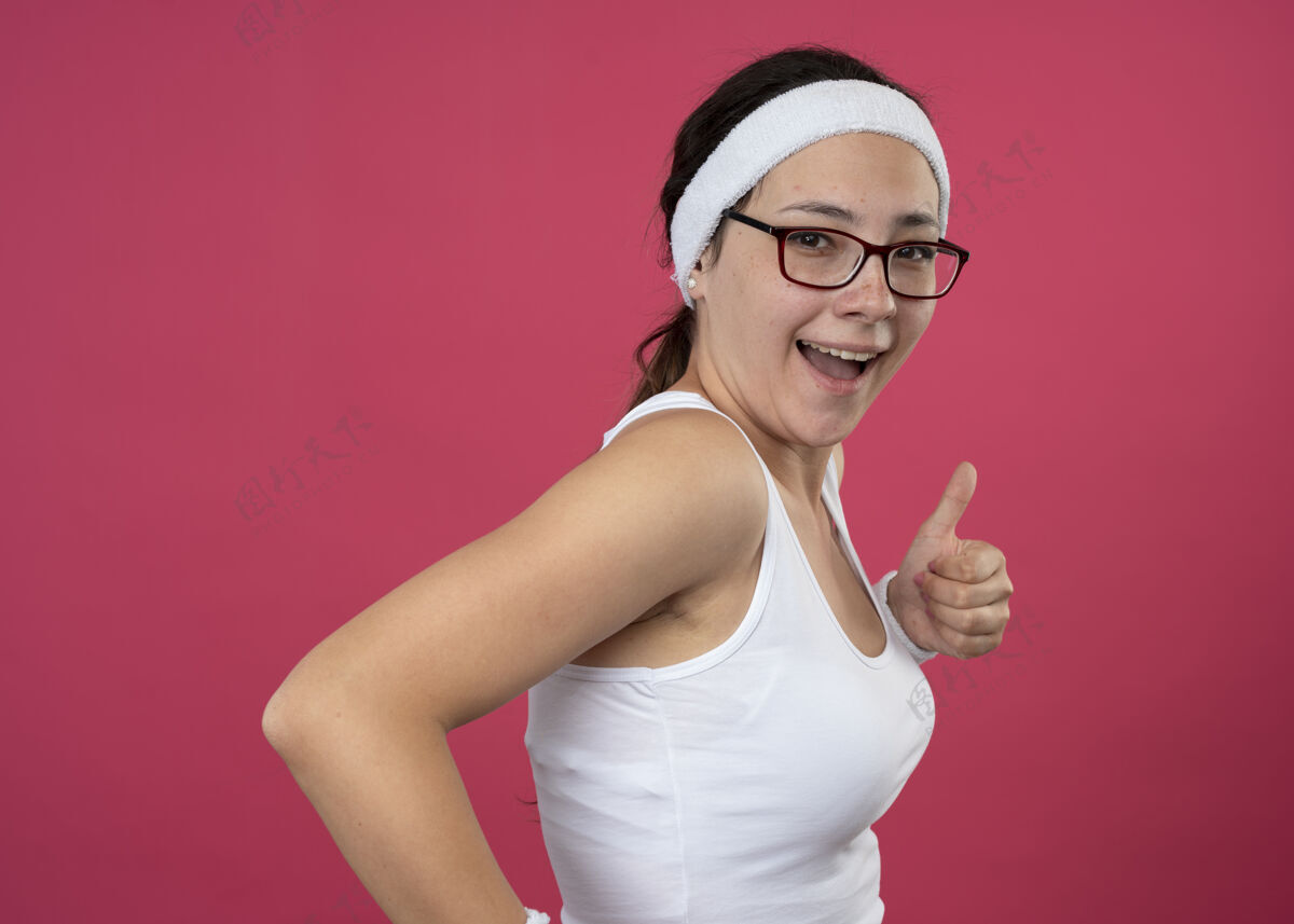 立场戴着眼镜 戴着头带和腕带的快乐的年轻运动女性站在一旁 竖起大拇指孤立地站在粉红色的墙上人运动腕带