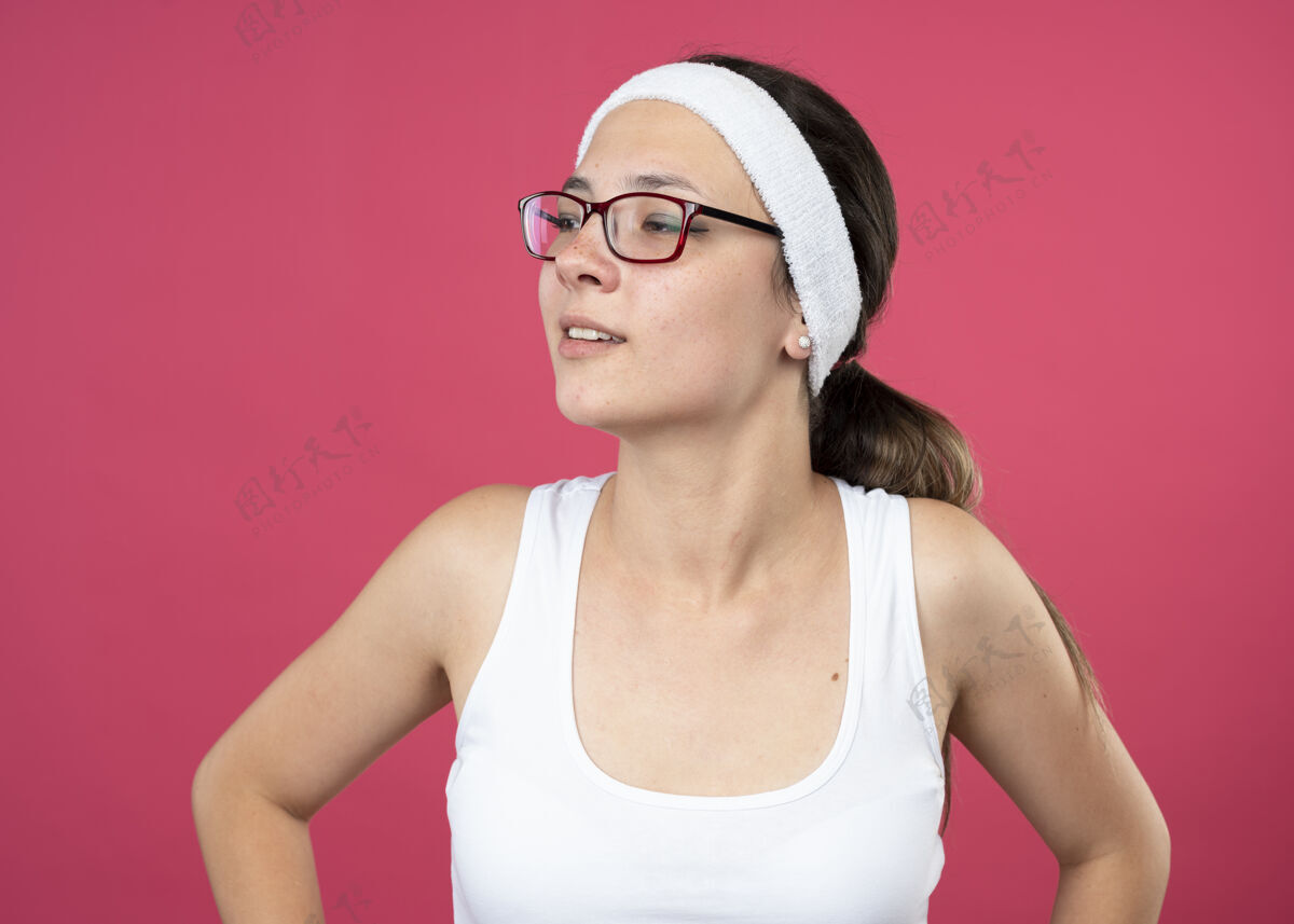 年轻戴着眼镜 戴着头带和腕带的快乐的年轻运动女性看着隔离在粉红色墙上的一面壁板女孩头带