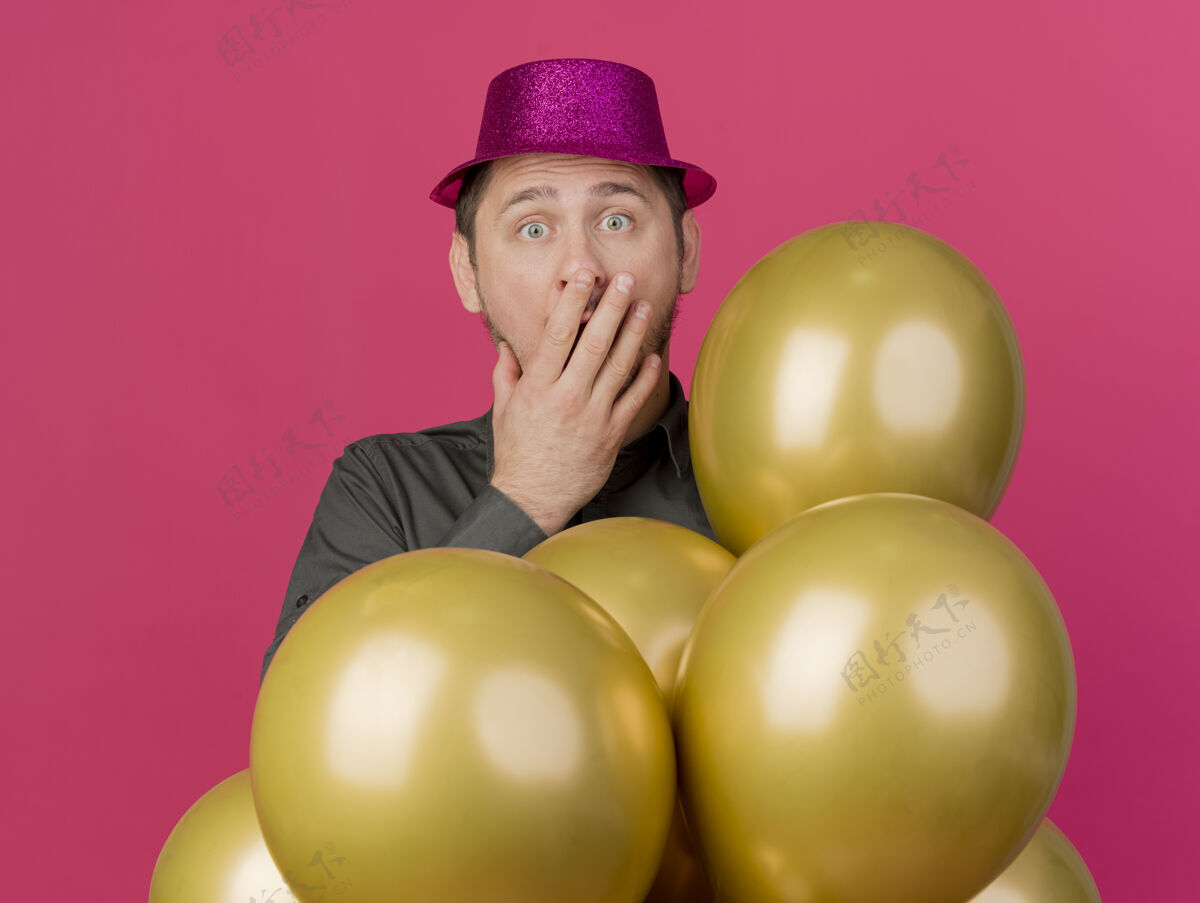气球害怕的年轻人戴着粉红色的帽子站在气球后面 把嘴孤立在粉红色站着害怕穿上