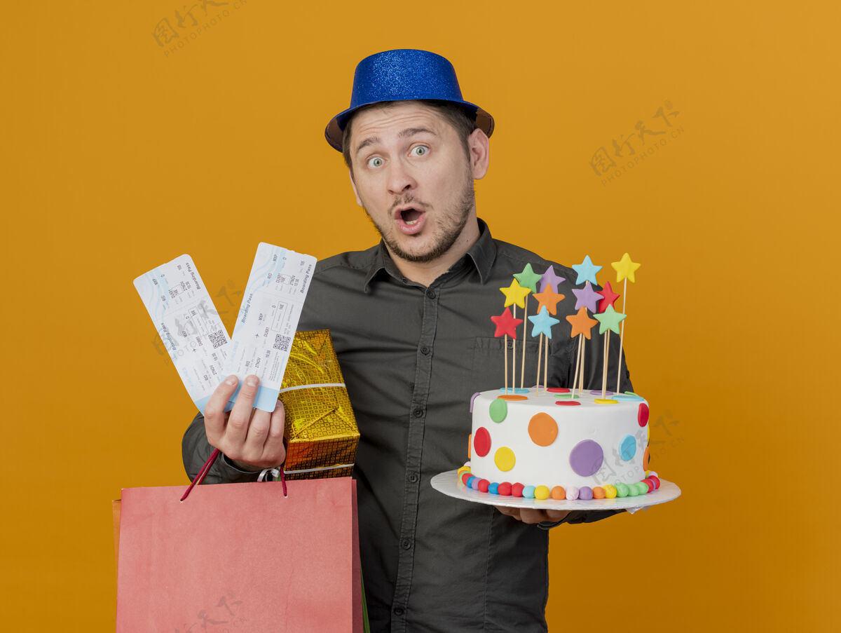 蓝色一个戴着蓝色帽子的年轻人拿着礼物 蛋糕和门票被隔离在橙色的墙上礼物惊喜帽子