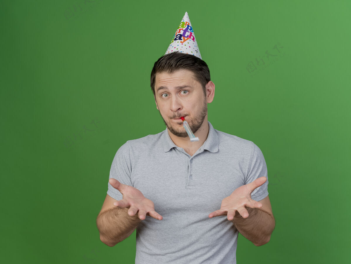 手困惑的年轻人戴着生日帽吹口哨 摊开双手孤立在绿色生日家伙吹
