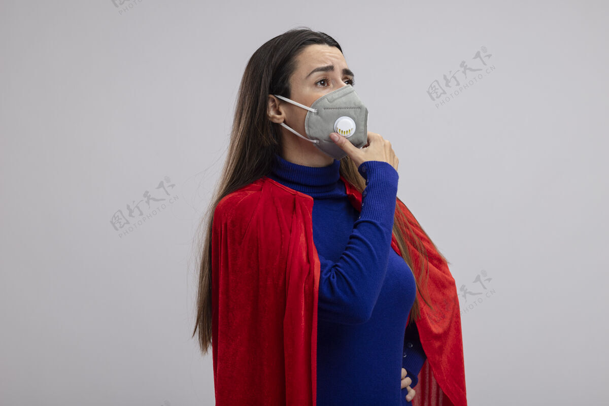 穿坚强的年轻超人女孩戴着医用口罩看着侧面 抓住下巴隔离在白色上思考壁板下巴