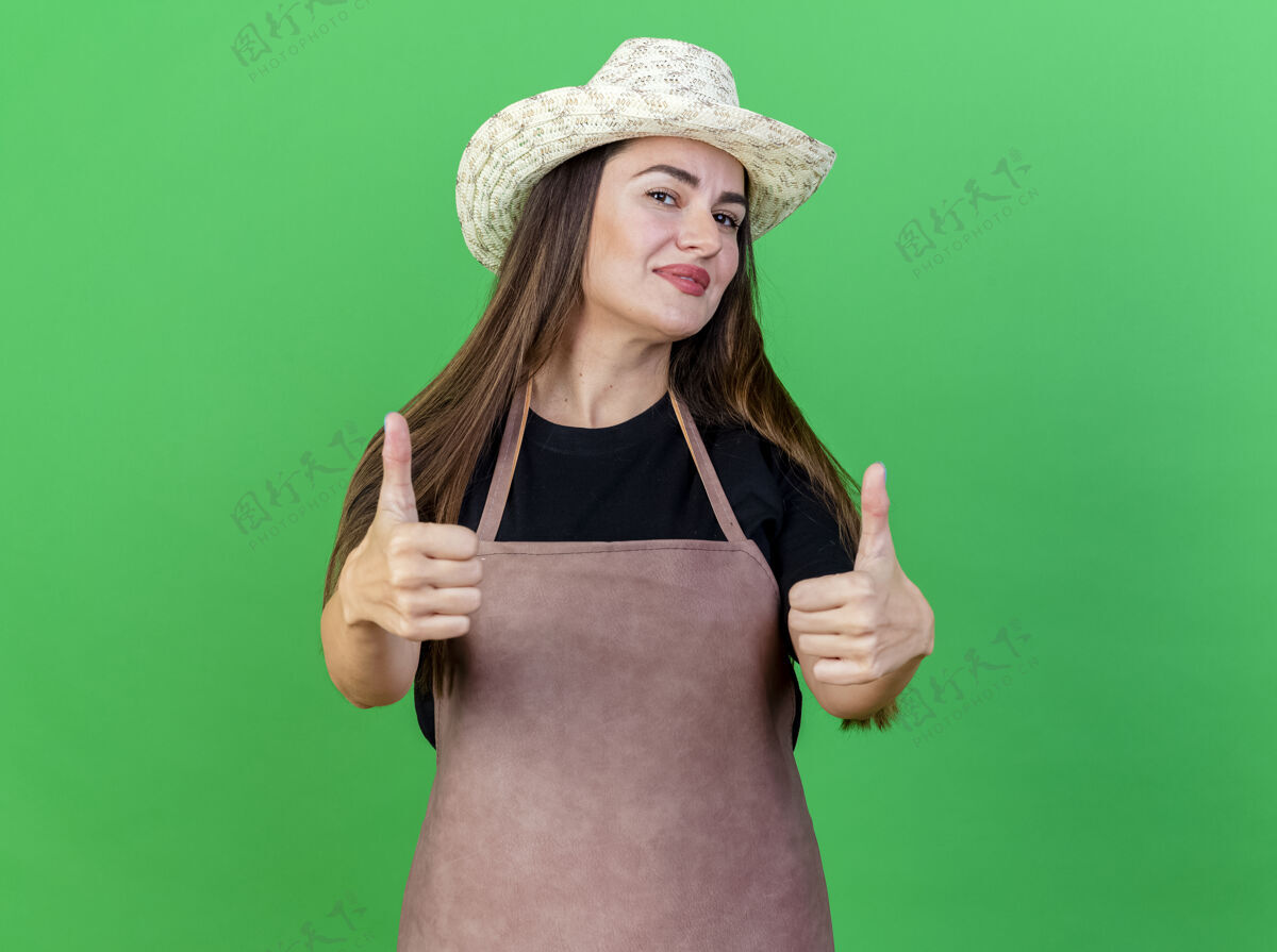 穿一个穿着制服的漂亮园丁女孩戴着园艺帽 在绿色的草坪上竖起大拇指请拇指表演