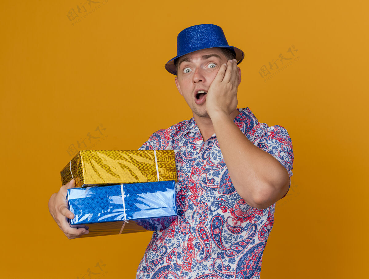 男人害怕的年轻人戴着蓝色帽子拿着礼品盒把手放在脸颊上孤立的橙色年轻橙色蓝色