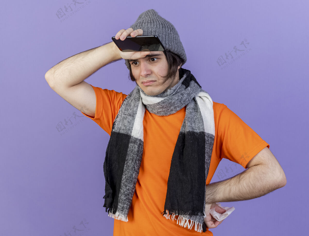 疾病悲伤的年轻人戴着冬天的帽子 戴着围巾 手里拿着电话 手放在额头上 臀部孤立在紫色上悲伤围巾年轻