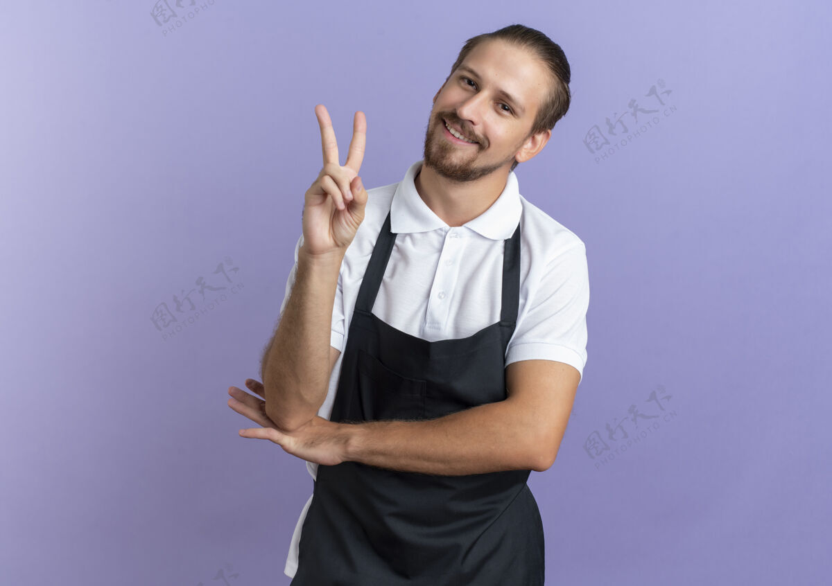 脸面带微笑的年轻英俊的理发师穿着制服做和平的标志 把手放在手肘下孤立在紫色的墙上和平帅气穿