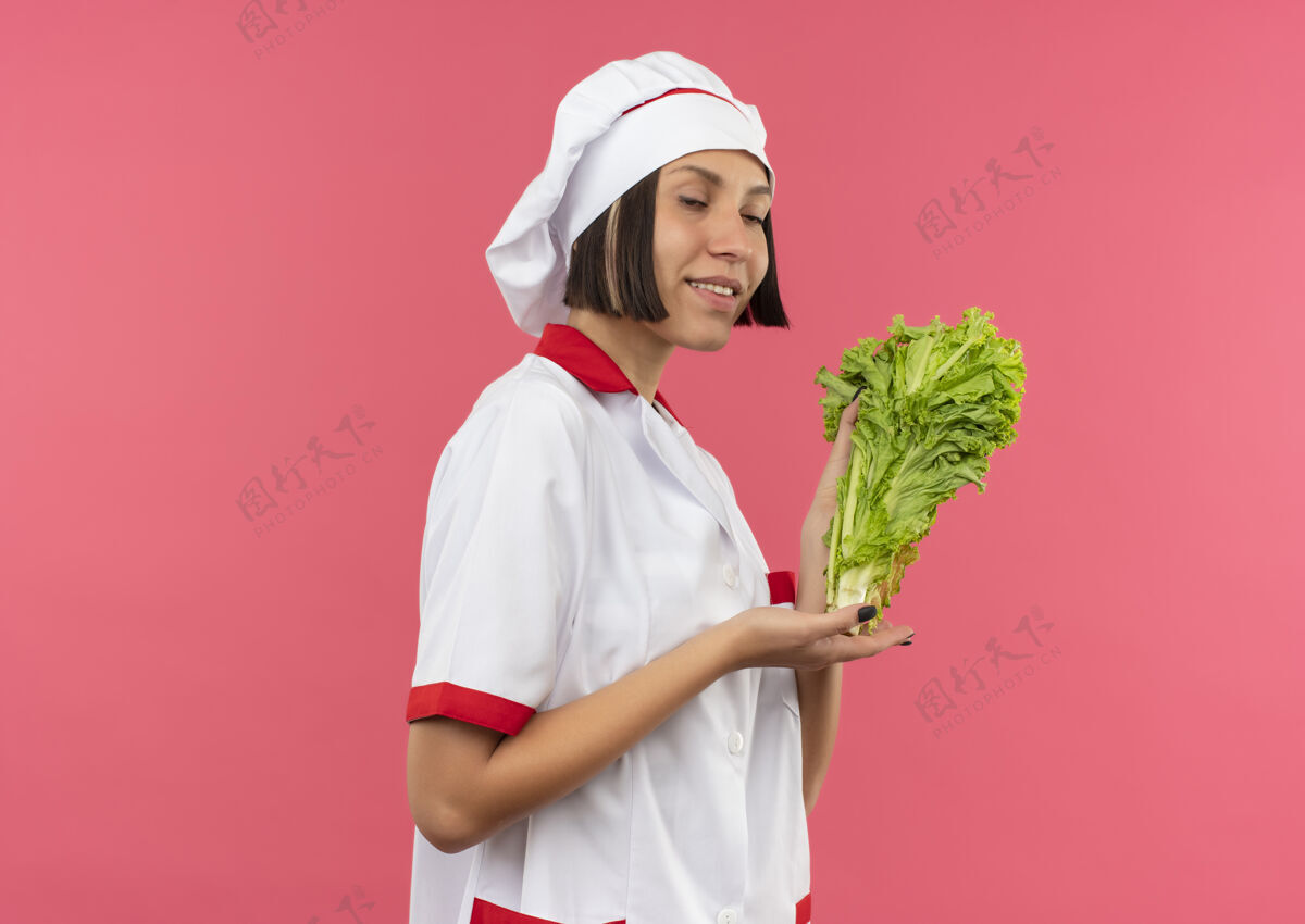 姿势身着厨师制服的年轻女厨师微笑着站在侧视图中 手拿莴苣 手指着隔离在粉红色墙上的莴苣女性指着拿着