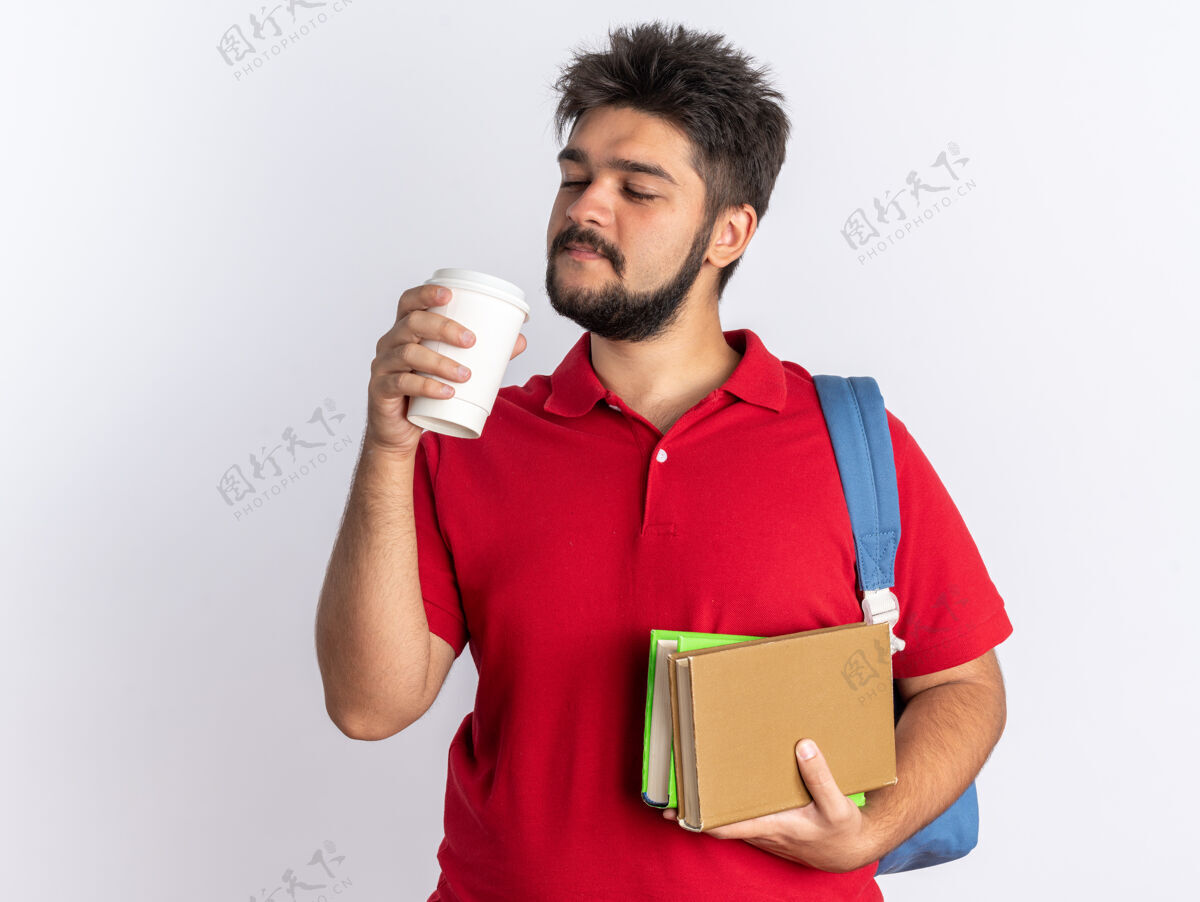 拿着年轻的留着胡子的学生 穿着红色马球衫 背着背包 手里拿着笔记本和咖啡杯 站在白色的背景下 看上去自信地微笑着笔记本胡子咖啡