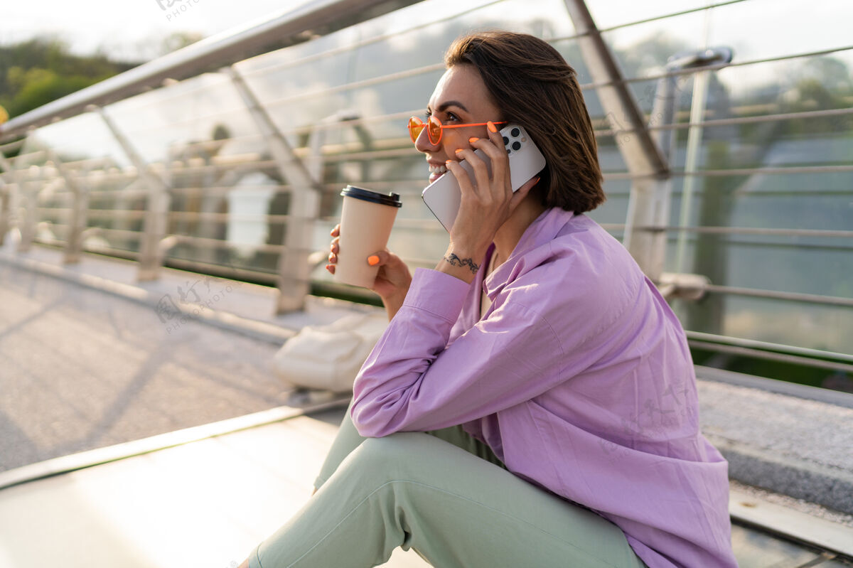 美丽时尚短发女子坐在摩登桥上 一边喝咖啡一边用手机夏天模型电话
