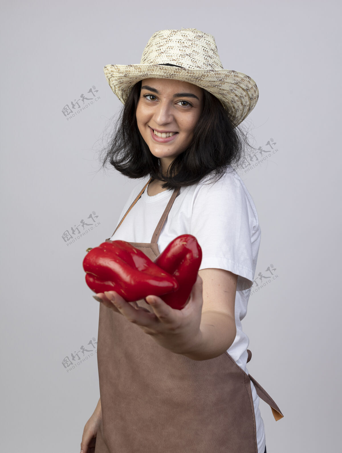 人微笑着的年轻黑发女园丁穿着制服 戴着园艺帽 手里拿着隔离在白墙上的红辣椒黑发制服抱着