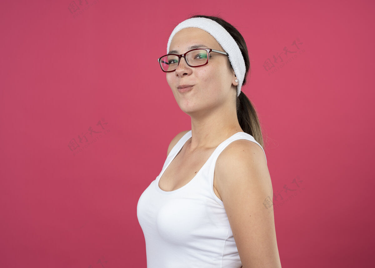 女人戴着眼镜 戴着头带和腕带的快乐的年轻运动女性站在一边 看着粉色墙壁上孤立的前方人侧面请