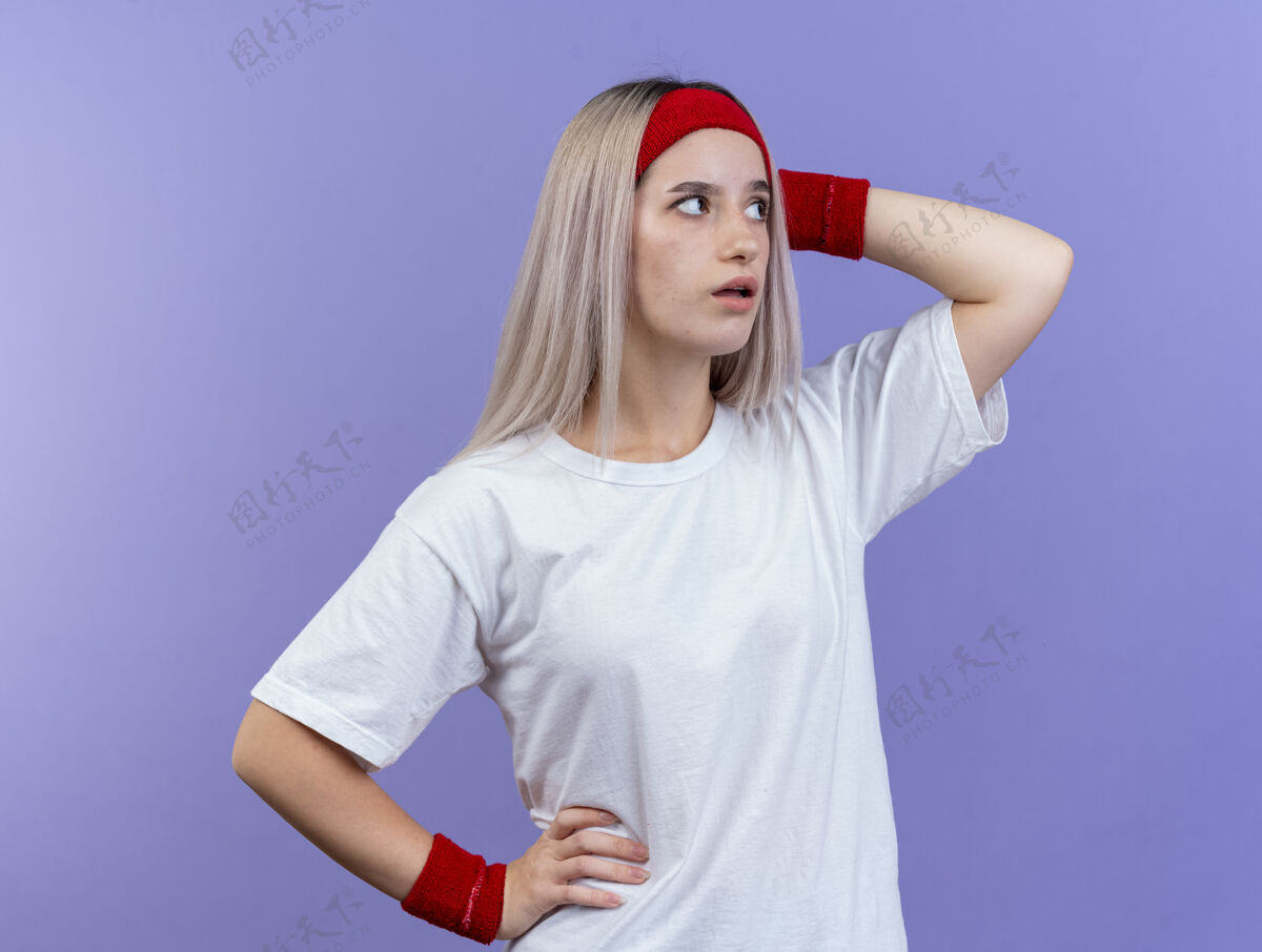 头焦虑的年轻运动女性戴着护腕戴着头带 手放在头上 看着紫色墙上孤立的一面人人佩戴