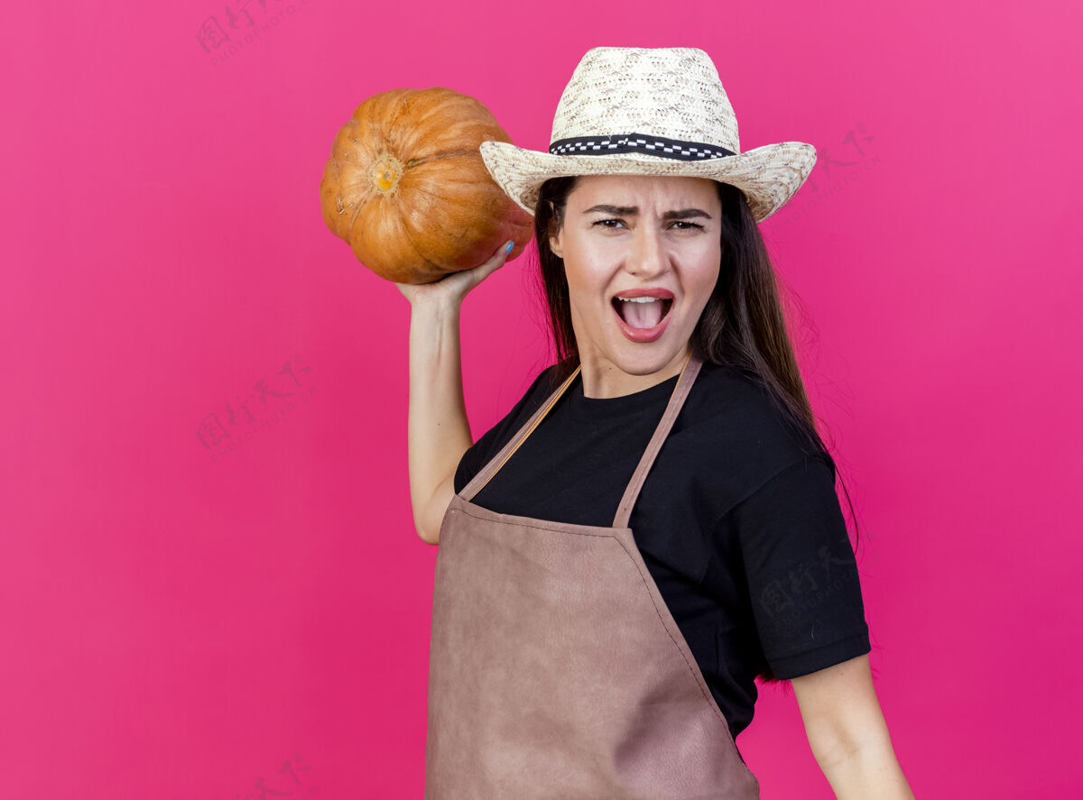帽子愤怒美丽的园丁女孩穿着制服 戴着园艺帽 手里拿着南瓜 背上孤立的粉红色举行花园背部