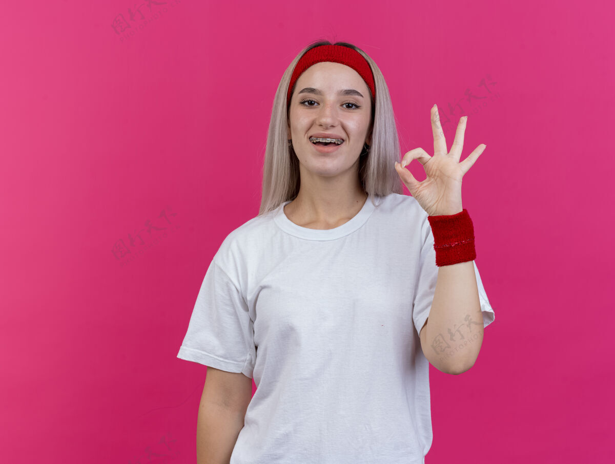 手势快乐的年轻运动女性戴着牙套戴着头带和腕带手势ok手势隔离在粉红色的墙上女孩女人腕带