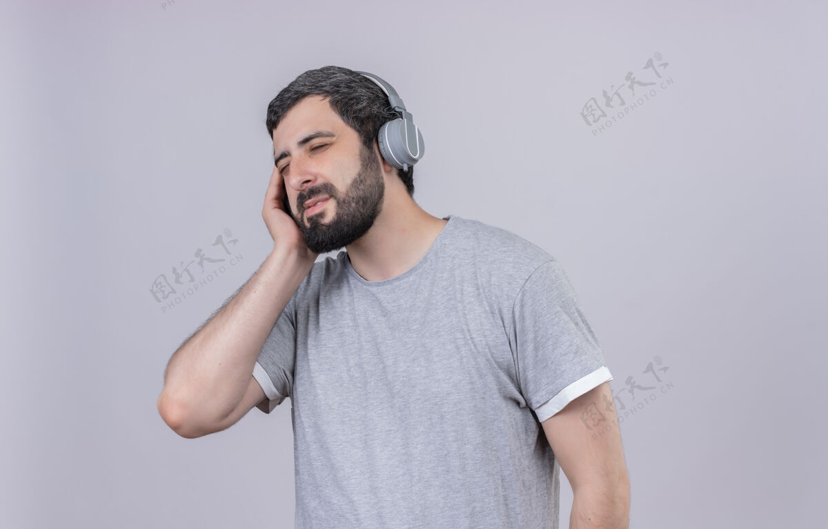 穿着高兴的年轻帅哥戴着耳机听音乐手戴着耳机闭着眼睛隔离在白墙上耳机脸感情