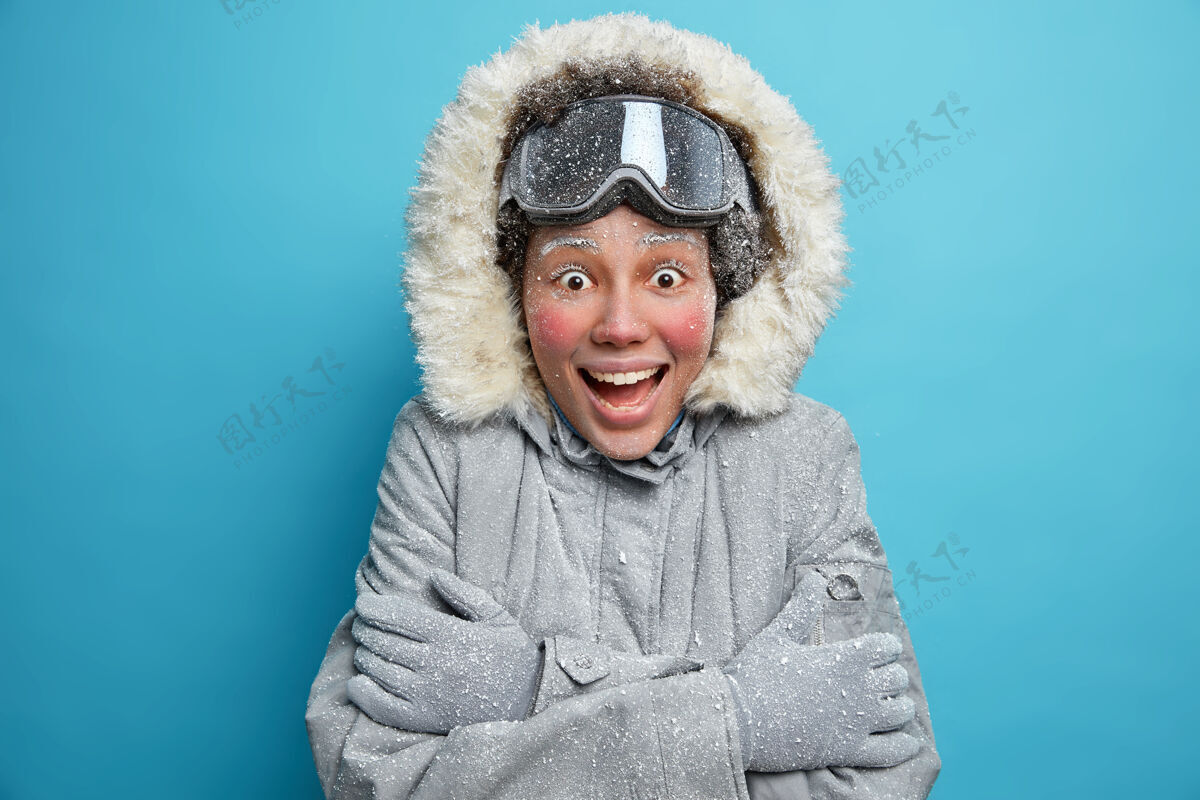 天气寒冷的冬季天气兴奋欢快的民族年轻女子 冻红了脸 在寒冷的天气里拥抱自己取暖 穿着保暖夹克和滑雪板护目镜 对一些情绪有反应雪寒假圣诞节