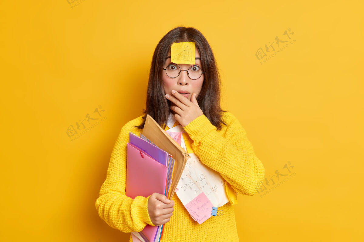 室内困惑惊讶的亚洲女学生的照片有备忘录贴在额头上准备课程作业携带文件夹和文件使教育项目在她的作业工作远程学习站立提醒通知