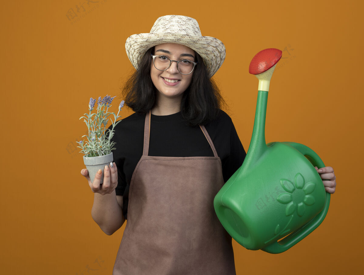浇水微笑着的年轻黑发女园丁 戴着眼镜 穿着制服 戴着园艺帽 拿着花盆和浇水罐 隔离在橙色的墙上 留着复制空间年轻人光学花园