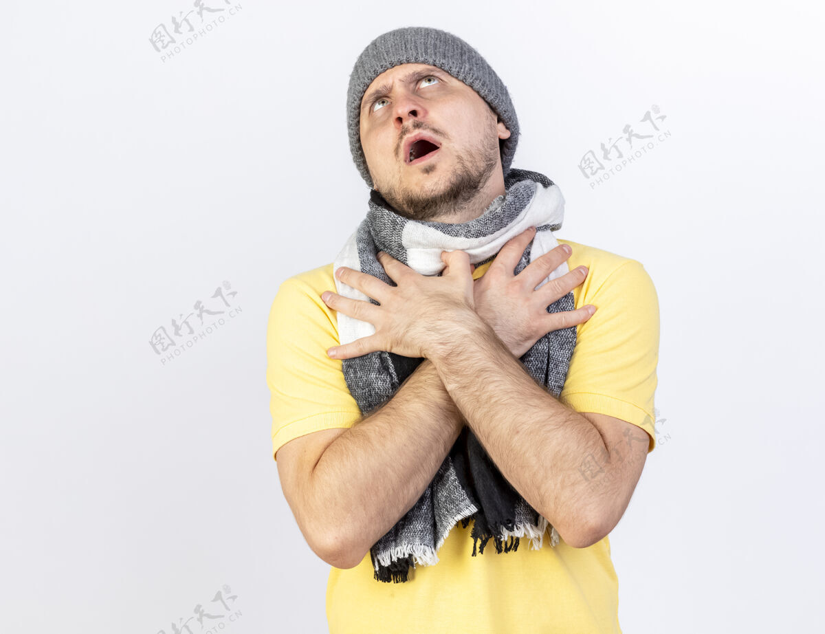 男人无知的年轻金发病斯拉夫男子戴着冬季帽子和围巾把双手放在胸前看着孤立的白色墙壁与复制空间围巾金发无知