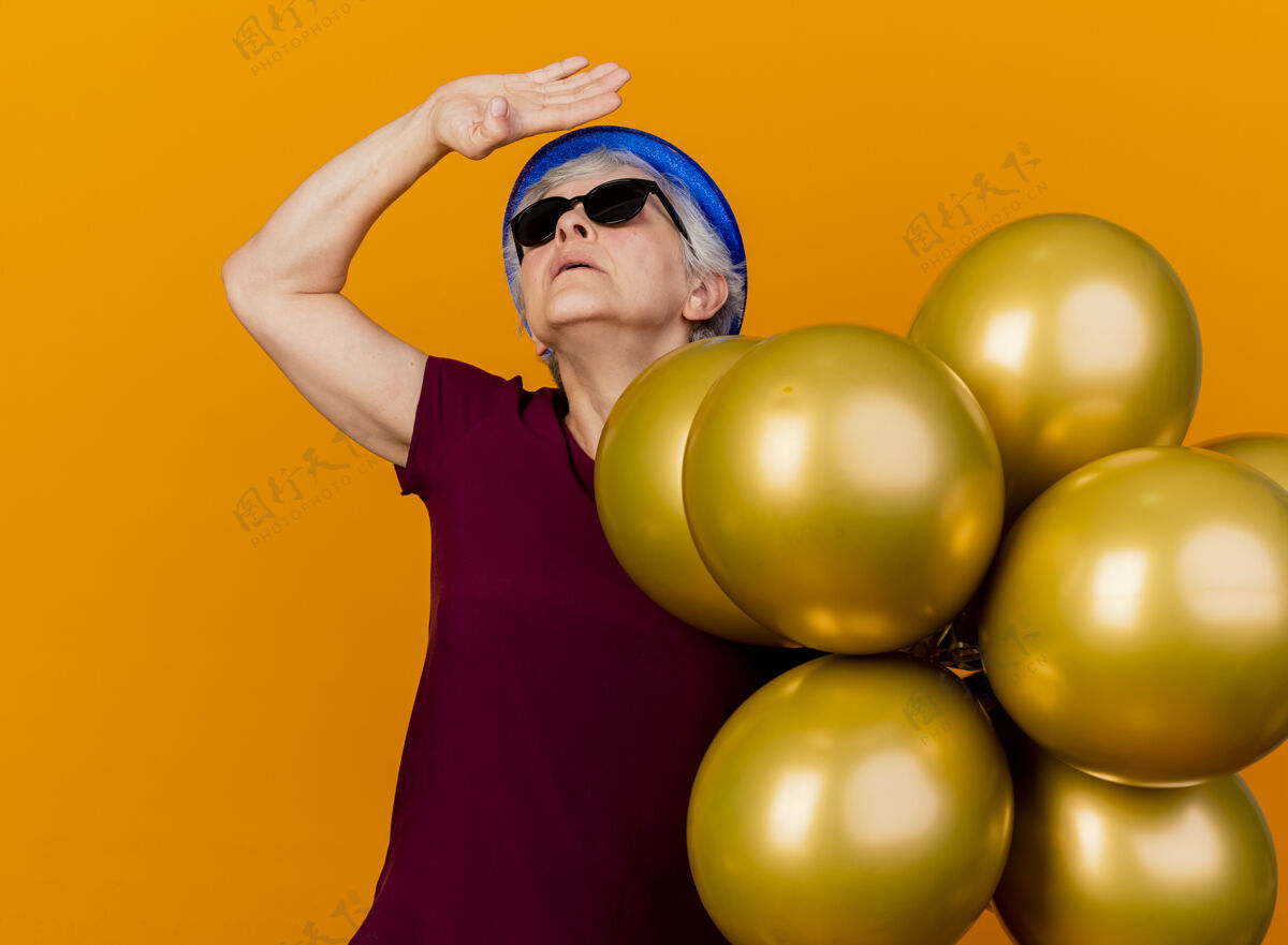 站着戴着派对帽 戴着太阳眼镜的自信的老妇人站在那里 手里举着氦气球 孤零零地仰望着橘黄色的墙上 还有复制空间戴着帽子气球