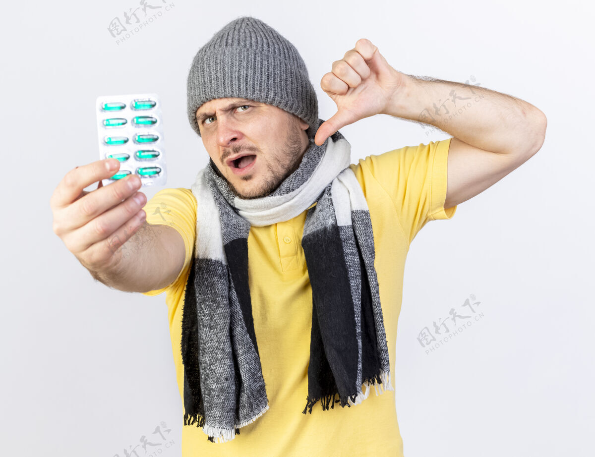 年轻人年轻的金发病斯拉夫男子戴着冬天的帽子和围巾拇指向下 持有一包医药丸隔离在白色的墙壁与复制空间男人金发围巾