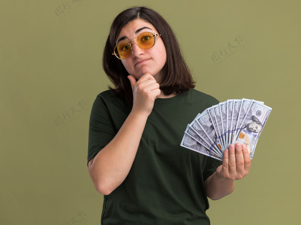下巴困惑的年轻漂亮的高加索女孩戴着太阳镜把手放在下巴上 把钱放在橄榄绿上迷茫绿眼镜