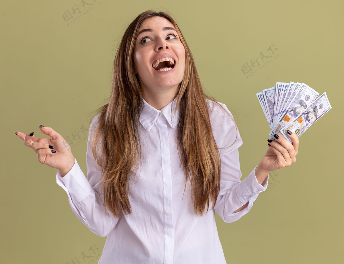 漂亮快乐的年轻漂亮的白人女孩拿着钱看着并指着橄榄绿的一边金钱橄榄指尖