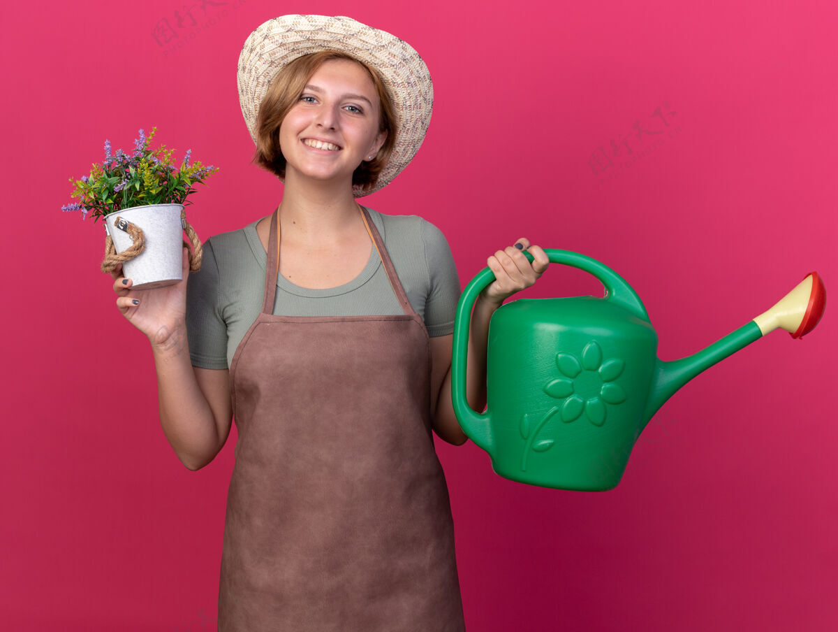 花盆面带微笑的年轻斯拉夫女园丁戴着园艺帽 手里拿着浇水罐 花盆里放着粉红色的花斯拉夫年轻女性