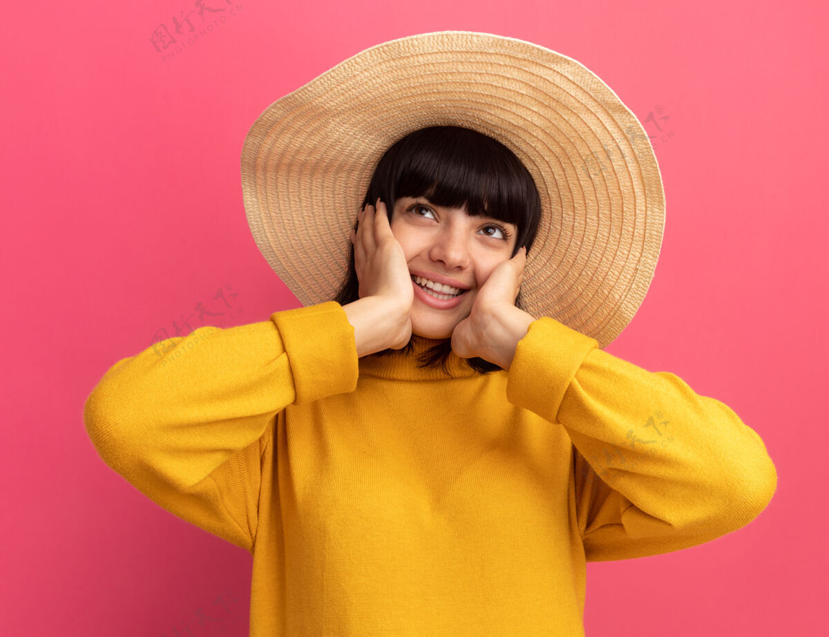 壁板高兴的年轻黑发白人女孩戴着沙滩帽把手放在脸上 看着粉红色的一面海滩手帽子