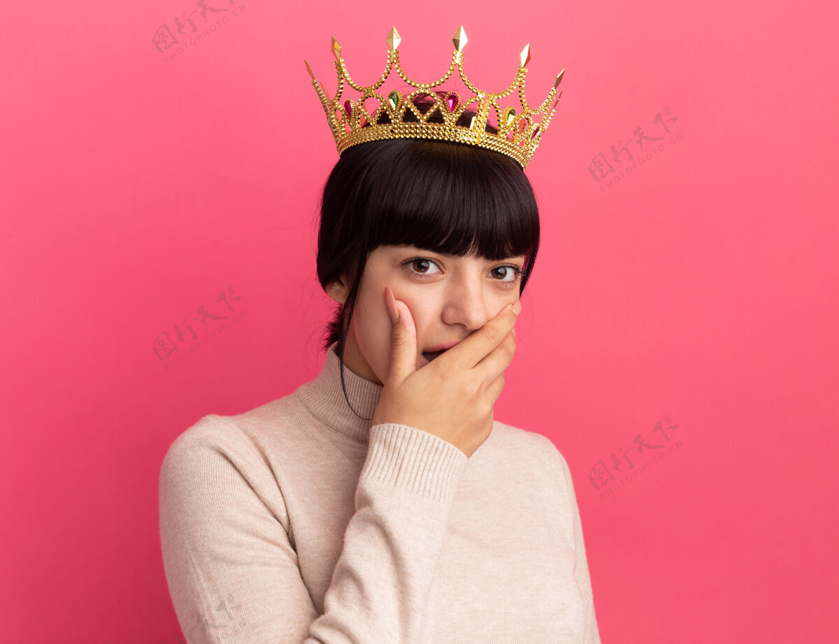 黑发震惊的年轻黑发白人女孩戴着皇冠把手放在嘴上 看着粉红色的相机手王冠毛衣