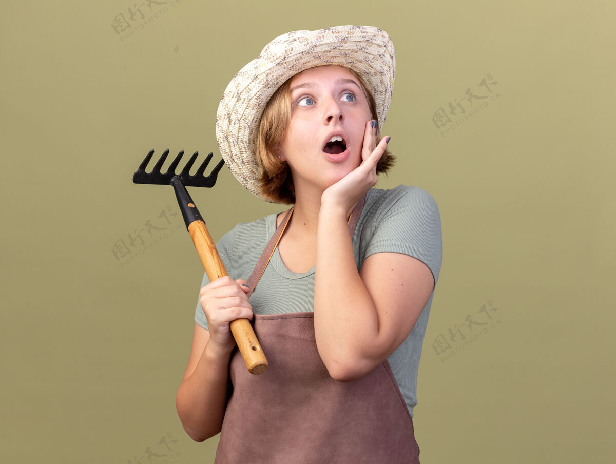 园艺兴奋的年轻斯拉夫女园丁戴着园艺帽把手放在脸上 拿着耙子看着橄榄绿的一面壁板女性帽子