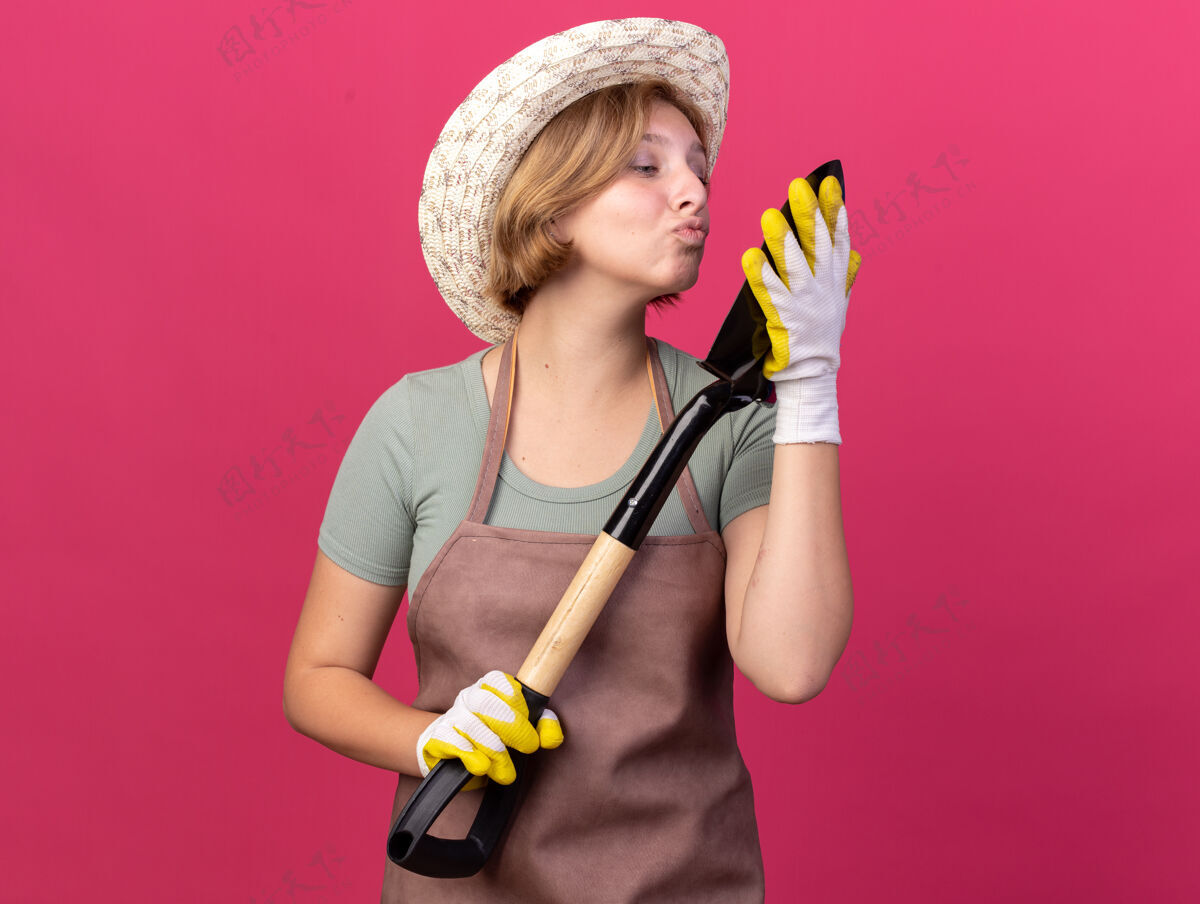 请高兴的年轻的斯拉夫女园丁戴着园艺帽和手套拿着并假装亲吻粉红色的黑桃黑桃女性园艺