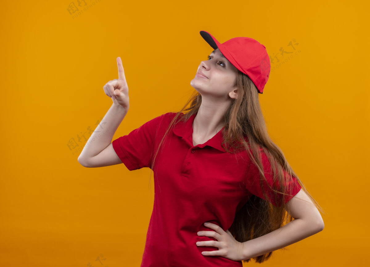 年轻身着红色制服的年轻送货女孩高兴地指着并抬起头 手放在腰上孤立的橙色空间腰部指向向上